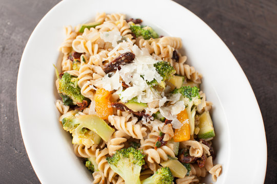  
Large White Bowl of cooked pasta topped with sundried tomatoes, bell peppers, broccoli, and parmesan cheese slightly above shot
