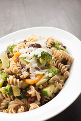  
Large White Bowl of cooked pasta topped with sundried tomatoes, bell peppers, broccoli, and parmesan cheese close up shot
