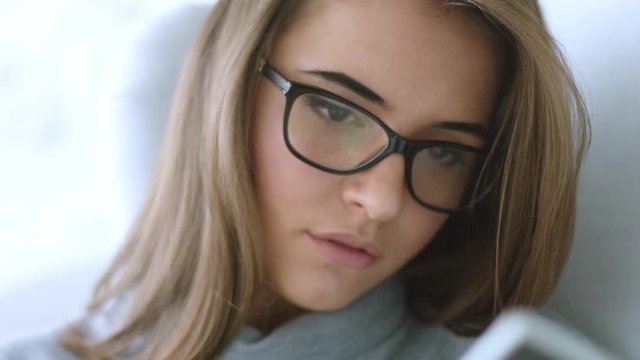 Close up view of attractive young brunette girl in glasses relaxing at home on the armchair reading a book