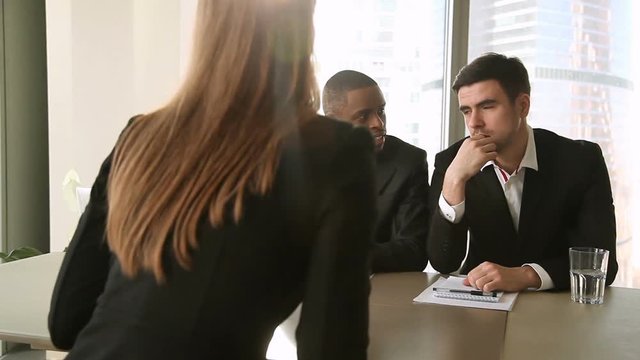 Multicultural businessmen wearing suits interviewing young lady, searching for a job, listening to female applicant telling about working experience, reviewing resume, asking answering questions