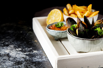 Mussels with herbs in a bowl with lemon and French fries on a white wooden board. Seafood. Food at the shore of the French Sea. Dark background.