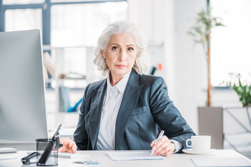 Confident senior businesswoman sitting at workplace and writing with pen