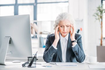 Obraz premium Senior businesswoman with headache sitting at workplace with desktop computer