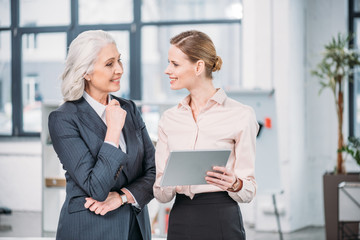 Two smiling businesswomen using digital tablet and talking in office
