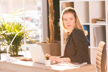 Beautiful young businesswoman sitting at desk with laptop and looking away