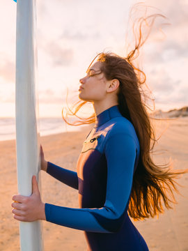 Beautiful surf girl with long hair holding surfboard at sunset.