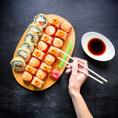 Sushi rolls on wood board, sauce, ginger, wasabi and woman hand holding chopsticks on dark background. Top view. Flat lay.