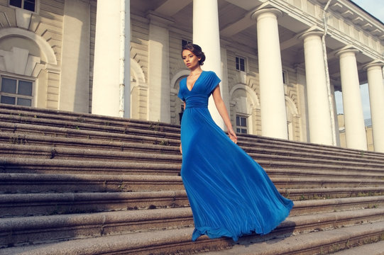 Fashion style portrait. Young elegant woman in blue long flying dress posing at stairway against old city building