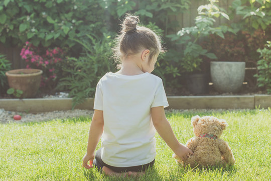 Toned photo of little girl holding her soft toy sitting on the grass with her back, selective focus