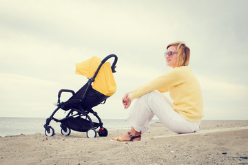 Smiling mother sitting on beach by the sea next sleeping baby in stroller
