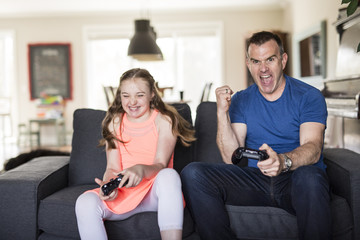 Obraz premium Handsome father and his cute little daughter are playing game console and smiling while sitting on couch at home.