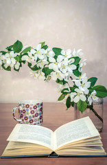 An open book on the table with a branch of apple blossoms and a cup of tea