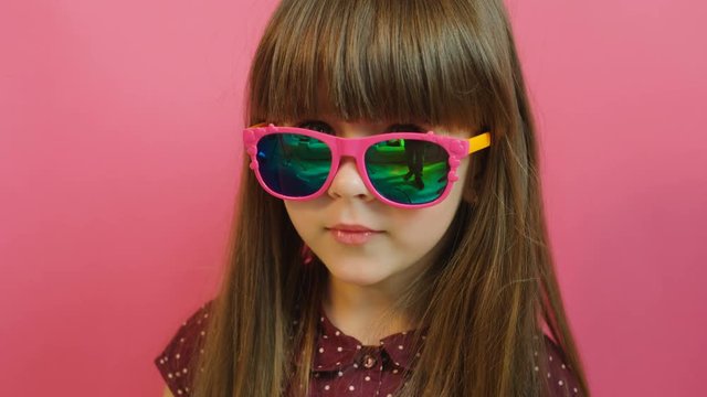 Face portrait of cute little girl in pink glasses looking at the camera. Pink background. Close up
