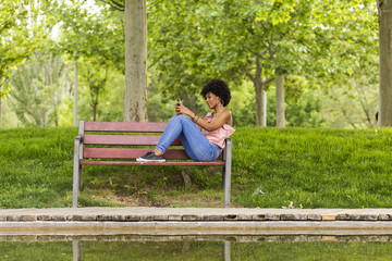 Fototapeta premium portrait of a Happy young beautiful afro american woman sitting on a bench and listening to music on her mobile phone. Green background. Lifestyles. Casual clothing.