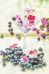 Glass goblet  with a refreshing drink, around many different berries blueberries and blueberries and ice cubes. Summer holiday mood. A light photo with a soft selective focus.
