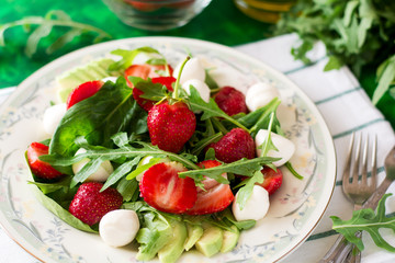 Fresh vegetarian salad with spinach, arugula, avocado slices, strawberries and mini mozzarella on green wooden table. Selective focus