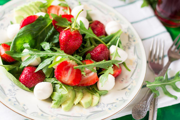 Obraz na płótnie Canvas Fresh vegetarian salad with spinach, arugula, avocado slices, strawberries and mini mozzarella on green wooden table. Selective focus
