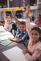 Portrait of happy schoolkids raising their hands in classroom