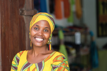 Young African woman in traditional clothes standing in entrance of her store, smiling at camera - obrazy, fototapety, plakaty