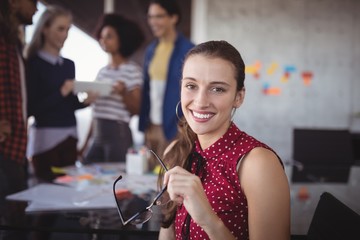 Smiling businesswoman holding eyeglasses with team in background
