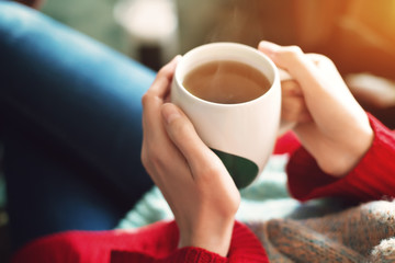Close up of pretty woman's hands in red sweater holding cup of tea in the morning sunlight.