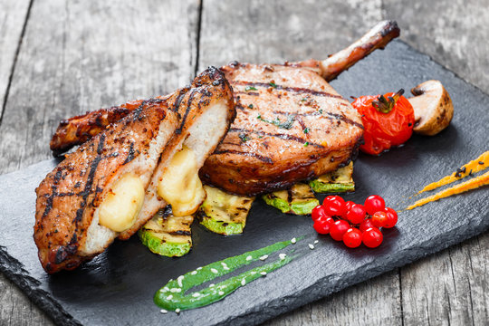 Roasted Pork steak on bone stuffed with cheese, grilled vegetables and berries on stone slate background on wooden background close up. Hot Meat Dishes. Top view with copy space