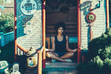 Young charming black girl is sitting cross-legged in Chinese pagoda and practicing relaxing yoga...