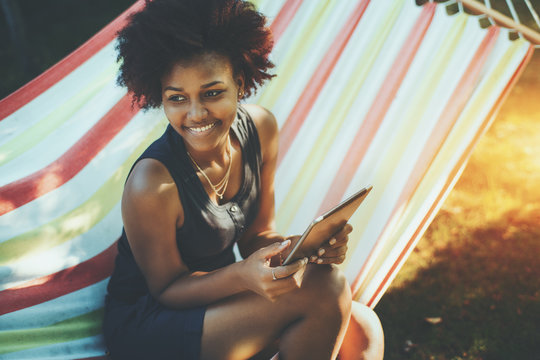 Laughing young cute afro american girl with curly hair is sitting and relaxing in colourful striped hammock, holding digital tablet and happily looking aside, sunny spring or summer day