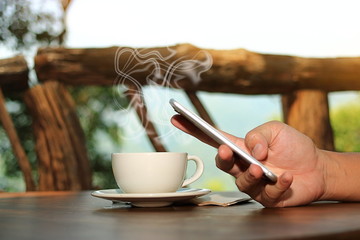 Close up coffee cup with hands of young man holding mobile phone in nature background. Selective focus and shallow depth of field.