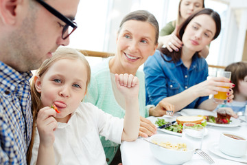 Little girl sitting on her father knees and showing tongue while eating fresh strawberry