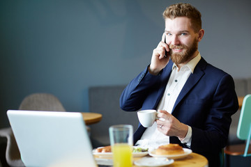 Portrait of handsome businessman speaking by phone and using laptop in cafe, looking at camera smiling charmingly