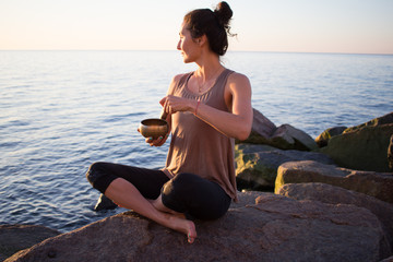 Fototapeta na wymiar young sporty woman play on Tibetan bowl and sitting on the stones, beautiful asian woman watching sunrise in the sea in yoga pose 