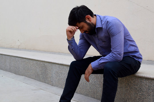 Stressed and sad young man sitting outside holding head with a hand looking down. Human emotion feelings, sad bearded man