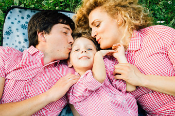 Happy family of three lying on the grass. Parents kissing them little son.