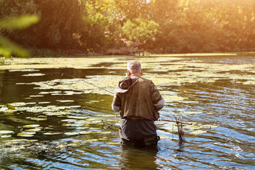 Old fisherman fishing in a lake or river with a fishing rod on a sunny day. Man standing in the water