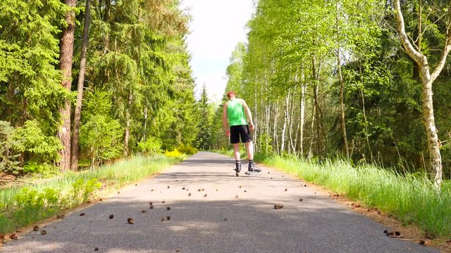 Rear view to inline skater in green running singlet . Outdoor inline skating on smooth asphal in the forest. Light skin man kick into pine cones on the road, moving with center of gravity.
