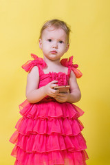 Girl toddler in a pink dress on a yellow background holding a smartphone and looking at the camera