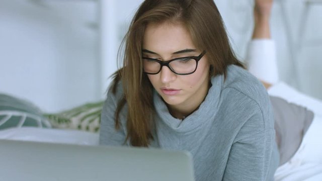 Beautiful girl in glasses relaxing on bed using laptop