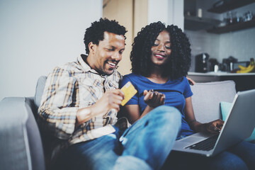 Smiling young african american couple shopping online through modern laptop using credit card at home. Horizontal,blurred background.