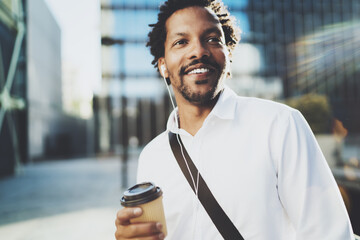 Smiling American African man in headphone walking at sunny city with take away coffee and enjoying to listen to music on his smartphone.Blurred background.Cropped.