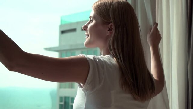 Woman in white t-shirt unveiling curtains and looking out of window. Enjoying the sea view outside