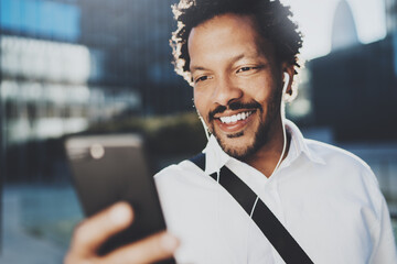 Smiling African American man in headphone making video call via mobile phone in hand.Concept of guy using Internet-enabled electronic device outdoor.Blurred background.