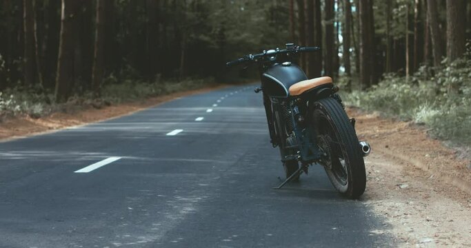 Back view of Caucasian man walking towards his custom built cafe racer motorcycle on a forest road. 4K UHD RAW edited footage 