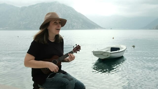 Young woman sits on beach, singing and playing guitar outdoors. Pretty woman with long hair, wearing hat, black T-shirt and jeans, sitting on rock, playing little guitar and singing funny song
