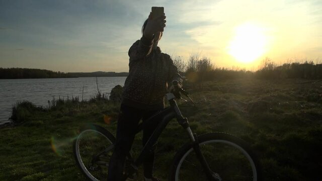 A young woman in a spotted hoody talking on a video call using her phone while holding a bicycle on a summer evening at sunset