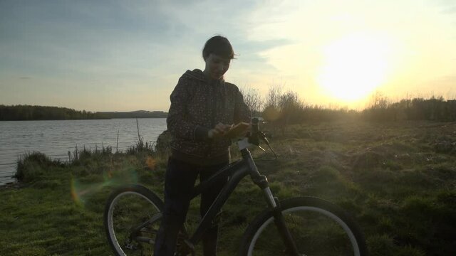 A young woman in a spotted sweatshirt uses her smartphone, then starts to rejoice and jump holding her bike on a summer evening at sunset