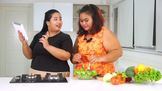 Video footage of two young overweight women preparing vegetable salad in the kitchen while looking at the recipe on a digital tablet