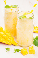Freshly blended yellow mango fruit smoothie in glass jars with straw, mint leaves, mango slices, close up. Soft white wooden board background.