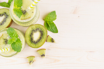 Fototapeta na wymiar Freshly blended green kiwi fruit smoothie in glass jars with straw, mint leaf, cute ripe berry, top view. White wooden board background, decorative border, copy space.