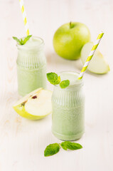 Freshly blended green apple fruit smoothie in glass jars with straw, mint leafs, apples. White wooden board background, copy space.